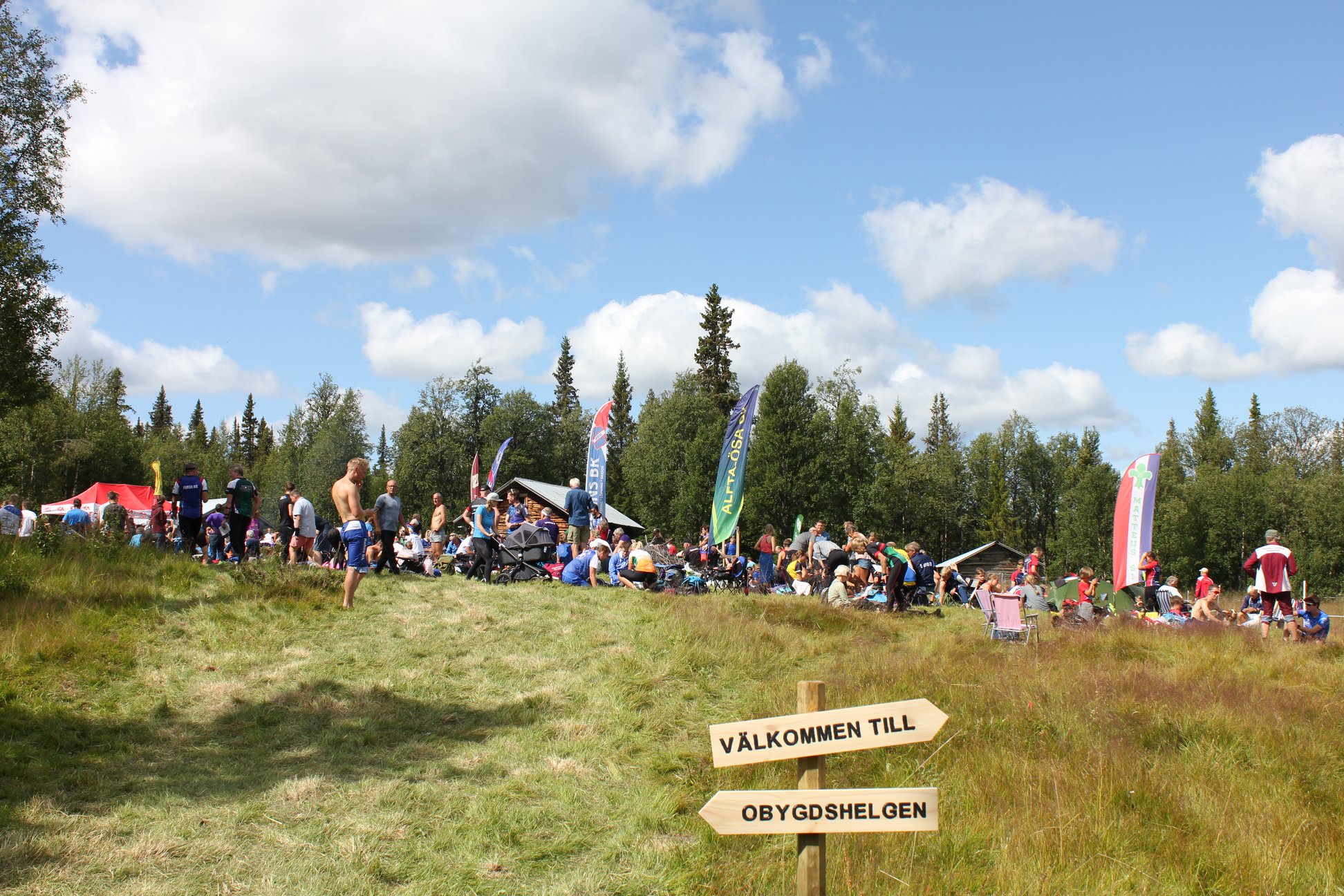 Äng med människor och vimplar och skylten "Välkommen till Obygdshelgen" i förgrunden
